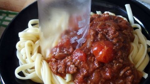 Ein großes Stück Spaghetti wird mit einem Küchenmesser in einem schwarzen Teller mit Tomatensauce kleingeschnitten.