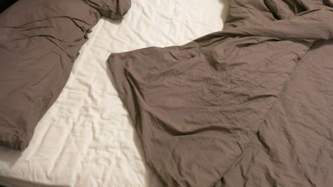 Auf der unordentlichen Bettmatratze liegen braune Bettlaken und eine abgedeckte Bettdecke in einem Schlafzimmer.