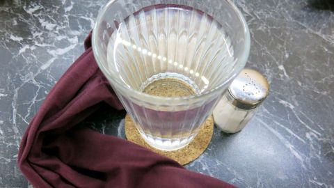 Ein Glas mit klarem Wasser steht auf einem Untersetzer, während ein rotbraunes Tuch daneben liegt, das für einen Salzwickel bei Halsschmerzen genutzt wird.