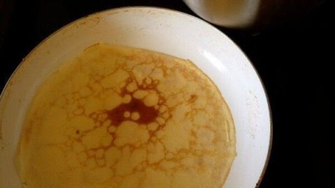 Eine goldbraune Pfannkuchen in einer Pfanne auf dem Herd, während im Hintergrund der Rührteig in einer Schüssel wartet.