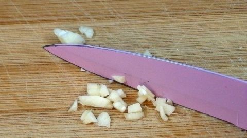 Frische Knoblauchstücke liegen neben einem pinken Messer auf einem Holzbrett, während es zerkleinert wird.
