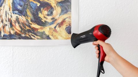Ein Föhn bläst warme Luft auf ein an der Wand hängendes abstraktes Poster, das anscheinend entfernt werden soll.