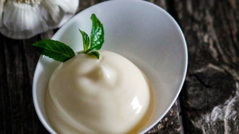 Um eine einfache Knoblauchmayo zu machen, pellen wir Knoblauch, pressen ihn mit der Knoblauchpresse und mischen ihn mit ganz normaler Mayo, und schon haben wier unsere Knoblauchmayo. Eine cremige Knoblauchmayo in einer weißen Schale steht auf rustikalem Holz, daneben liegt eine Knoblauchzehe.