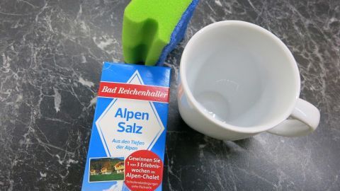 Ein Behälter mit Alpen Salz liegt neben einer leeren Tasse auf einer dunklen Oberfläche, bereit zur Reinigung von Schwarzteeresten.