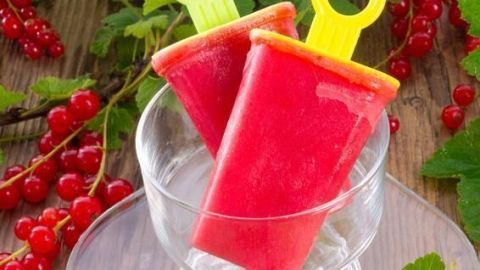 Zwei rote Wassereis mit bunten Stielen stehen in einer Glasschale, umgeben von frischen roten Johannisbeeren und grünen Blättern.