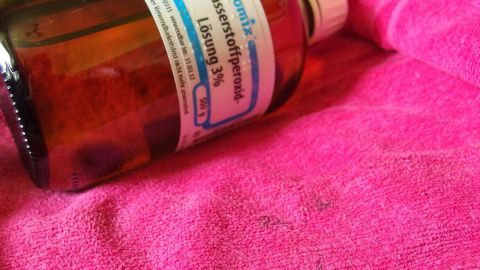 Meine Kinder essen sehr gerne Blaubeeren. Oft bleibt das nicht ohne Flecken. Diese entferne ich ganz einfach mit Wasserstoffperoxid. Eine Flasche Wasserstoffperoxid liegt auf einem pinken Tuch, bereit zur Anwendung gegen Blaubeerflecken.