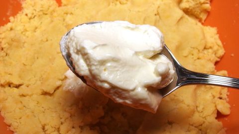 Mit etwas Quark oder Joghurt wird brösliger Mürbeteig wieder schön geschmeidig. Ein Löffel mit Sahne wird in einen Mürbeteig gehalten, der in einer orangefarbenen Schüssel liegt. Der Teig wirkt bröselig.
