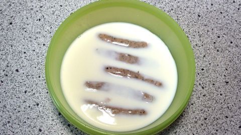 Knusprige Bratwürste liegen in einer grünen Schüssel, umgeben von Milch auf einer grauen Arbeitsplatte.