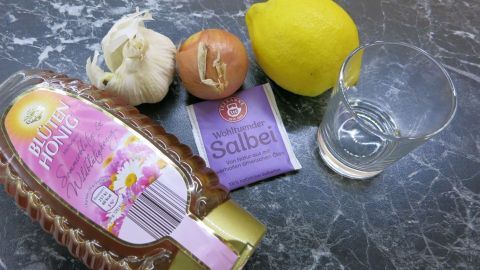 Honigflasche, Knoblauch, Zwiebel, Zitrone und leerer Glas auf grauem Tisch, daneben Salbeitee-Packung mit Text: "Wohlfühlender Salbei".