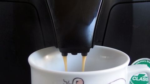 Einschlafprobleme wegen zu niedrigem Blutdruck? Kaffee mit Milch und etwas Zucker hilft. Dunkler Kaffee fließt in eine Tasse mit einer niedlichen Maus, während die Maschine im Hintergrund arbeitet.