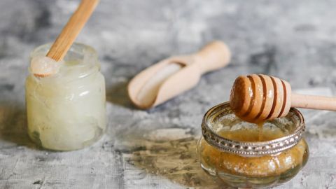 Ein Glas mit Lippenpeeling und ein Honiglöffel stehen auf einer neutralen Oberfläche, umgeben von einer ruhigen Umgebung.