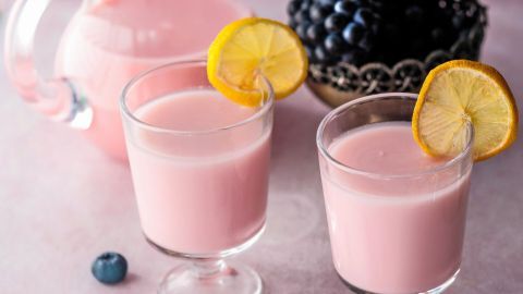 Zwei Gläser Heidelbeer-Milchshake stehen auf einem Tisch, umgeben von frischen Heidelbeeren und einem Krug mit Getränk.