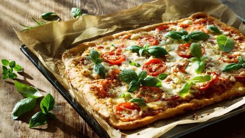 Eine frisch gebackene Pizza liegt auf Backpapier, belegt mit Tomaten und frischem Basilikum, auf einem Holztablett.