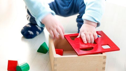 Ein kleines Kind sitzt auf dem Boden und versucht, ein rotes Formstück in eine Holzbox mit passenden Ausschnitten zu legen.