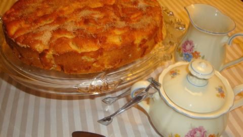 Ein saftiger Apfelkuchen steht in einer Glasplatte, umgeben von einer schönen Teekanne und einer Milchkanne auf einem gestreiften Tisch.