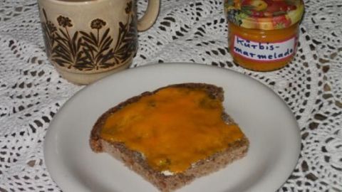 Auf einem Teller liegt ein Stück Brot mit Kürbismarmelade, daneben eine Tasse Kaffee und ein Glas Marmelade.