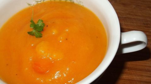 Eine cremige Kürbis-Karotten-Kartoffel-Suppe in einer weißen Tasse auf einem Holzuntergrund, mit einem grünen Kräuter-Topping.