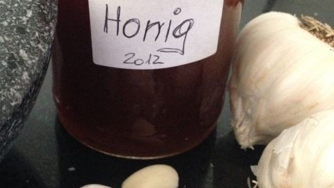 Ein Glas Honig steht auf einer Oberfläche, umgeben von frischem Knoblauch und einer Steinmörser, was auf ein Heilmittel hinweist.