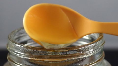 Ein Löffel mit Honig schwebt über einem offenen Glas, bereit zum Entnehmen, auf einem neutralen Hintergrund.
