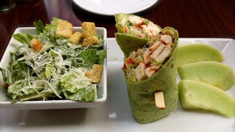 Eine grüne Wraps mit Hähnchen sind auf einem Teller neben einem Salat und melonenartigen Stücken angerichtet.