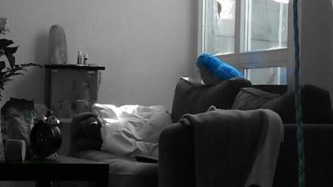 Person schläft auf Sofa, gestützt von Kissen, in lichtdurchflutetem Raum mit Pflanzen und Tisch im Vordergrund.