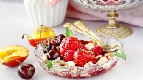 Ein farbenfrohes Obstarrangement zeigt frische Erdbeeren, Kirschen und Pfirsiche in einer eleganten Schale, umgeben von Blumen.