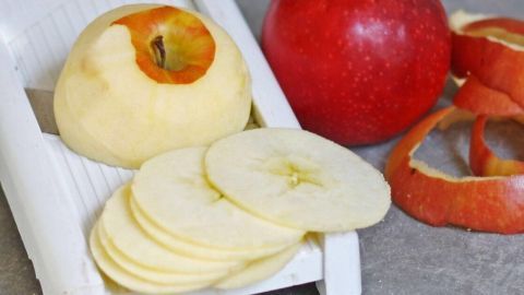 Scheiben von Äpfeln liegen auf einer weißen Hobel, während ein roter Apfel und Apfelschalen daneben liegen.