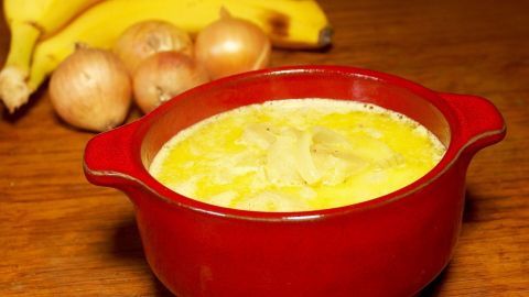 Eine rote Schüssel mit exotischer Bananen-Zwiebel-Suppe steht auf einem Holztisch, umgeben von Bananen und Zwiebeln.