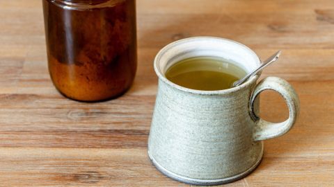 Eine graue Tasse mit Brühe steht auf einem Holztisch, neben einem Glasbehälter, der eine dunkle Flüssigkeit enthält.