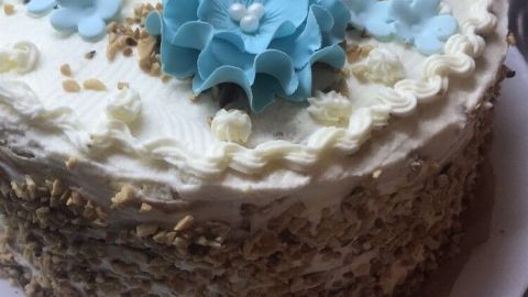 Eine dekorative Torte mit einer Cremehaube und blauen Blüten sitzt auf einem weißen Teller, umgeben von einem eleganten Ambiente.