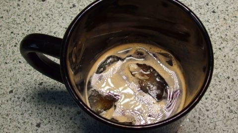 Eine Tasse Eiskaffee mit Eiswürfeln schwimmt in einer dunklen Flüssigkeit auf einer gemusterten Arbeitsfläche.