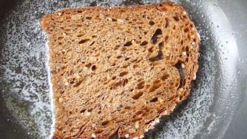 Graubrot in Butter knusprig braun anbraten, mit Salz bestreuen und genießen. In einer Pfanne brät ein Stück Brot in butterfarbener Flüssigkeit, umgeben von einem rustikalen Küchenambiente.