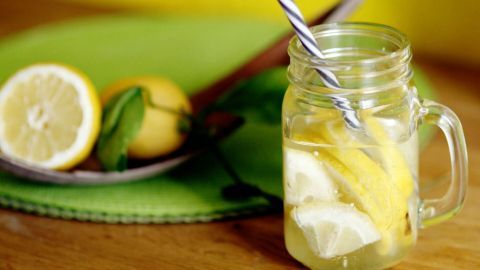 Ein Glas Limoncello mit Zitronenscheiben steht auf einem Holztisch, umgeben von frischen Zitronen und einem grünen Untersetzer.