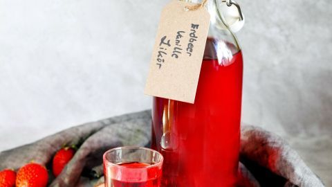 Ein Fläschchen Erdbeer-Vanille-Likör steht auf einem Holzbrett, umgeben von Erdbeeren und einem Glas Likör.