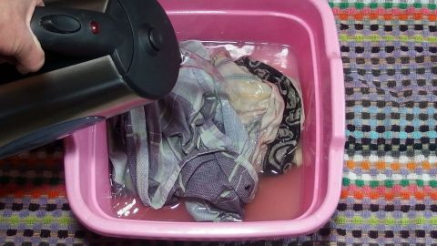 Ein Handgerät hält einen Wasserstrahl über bunte, kleine Mengen Kochwäsche in einer pinken Schüssel auf einem gestreiften Untergrund.