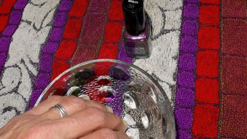 Ein Finger taucht in eine Schüssel, während ein Nagellack daneben auf einem bunten Tuch steht. Essig könnte zur Anwendung kommen.