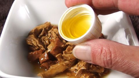 Eine Hand hält einen kleinen Behälter über eine Schüssel mit Katzenfutter, um eine Ergänzung für Haarballen zu geben.