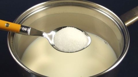 Eine Schöpfkelle mit Zucker schwebt über einem Topf mit Milch, während ein cremiger Pudding zubereitet wird.