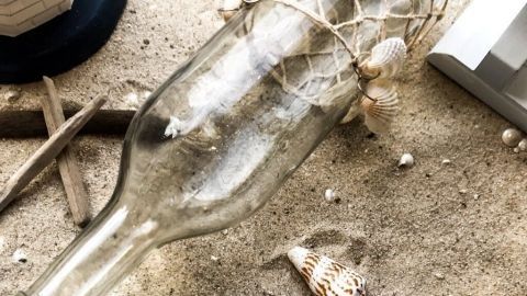 Eine dekorative Flasche liegt im Sand, umgeben von Muscheln, ideal für Dekosand-Projekte in maritimem Stil.