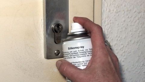 Eine Hand sprüht Schmiermittel in einen Schließzylinder an einer Tür, um dessen Funktion zu verbessern.