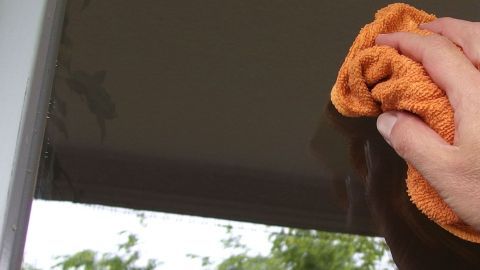 Eine Hand mit einem orangefarbenen Tuch reinigt ein Fenster, während Hintergrundgrün sichtbar ist, was auf einen Außenbereich hindeutet.