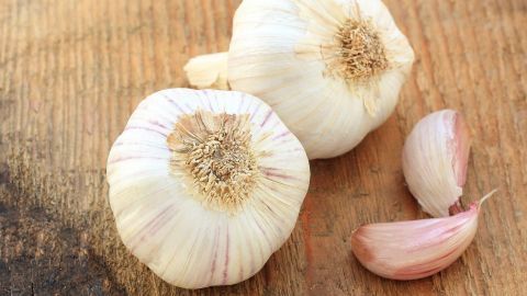 Zwei Ganze Knoblauchzehen liegen auf einem Holzbrett, während eine geschälte Knoblauchzehe daneben platziert ist.