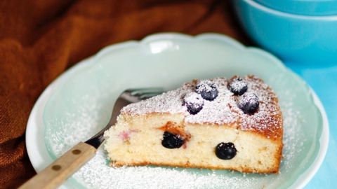 Geburtstagskuchen im Büro: Puderzucker mit einem Tee-Ei auf den Kuchen streuen. Funktioniert gut und man muss nicht sein ganzes Küchenequipment mitbringen. Ein Kuchenstück mit Puderzucker und Beeren auf einem Teller neben einer Schüssel mit Heidelbeeren steht auf einem Tisch.