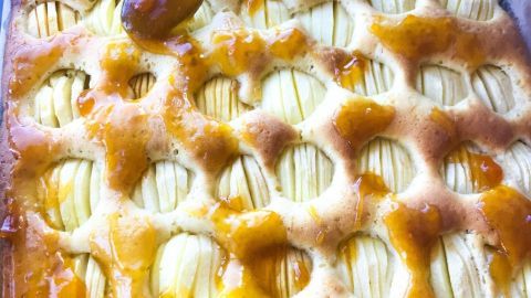 Ein handgehaltener Löffel verteilt Marmelade auf einem frisch gebackenen Blitz-Apfelkuchen in der Auflaufform.