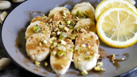 Scheiben von Bananen sind mit Pistazien und Zimt auf einem Teller angerichtet, umgeben von Zitronenscheiben und Nüssen.