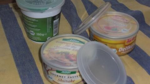 Drei Kunststoffdeckel bedecken Sahne- und Katzenfutterbecher auf einem gestreiften Tuch. Text: "Bio-Joghurt mild, 150g", "Allos Gourmet Pastete Shiitake", "Delikatessen".