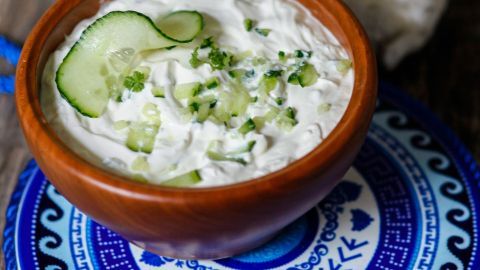 Ein Holzschale mit Tzatziki, garnierend mit Gurkenstücken, steht auf einem bunten, dekorativen Teller aus Keramik.