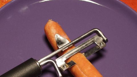 Manche Würstchen sind schwer abzupellen. Würstchen einfach mit dem Kartoffelschäler abschälen. Klappt super. Ein Wurstschäler entfernt die Haut von einem Würstchen auf einem lila Teller in einer alltäglichen Küchenumgebung.