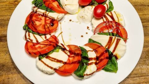 Auf einem weißen Teller liegt eine kunstvolle Anordnung aus Tomaten, Mozzarella und frischem Basilikum, garniert mit Balsamico.