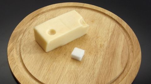 Ein Stück Käse liegt auf einem Holzbrett neben einem Würfel Zucker, der zur Frischhaltung verwendet wird.